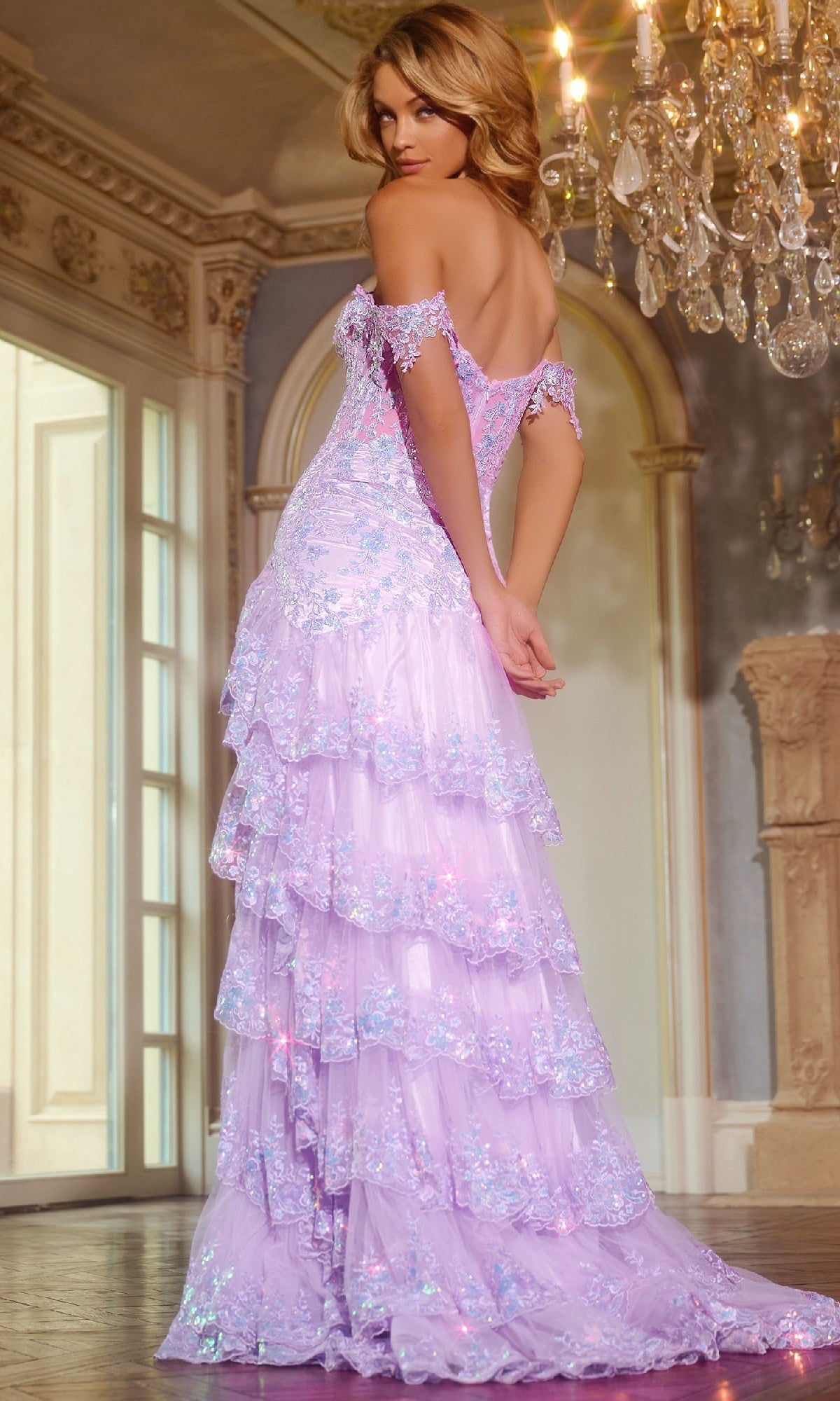 A woman wearing the Jovani 40397 Off-the-Shoulder Ruffle Prom Gown stands indoors near an ornate fireplace and chandelier, glancing over her shoulder.
